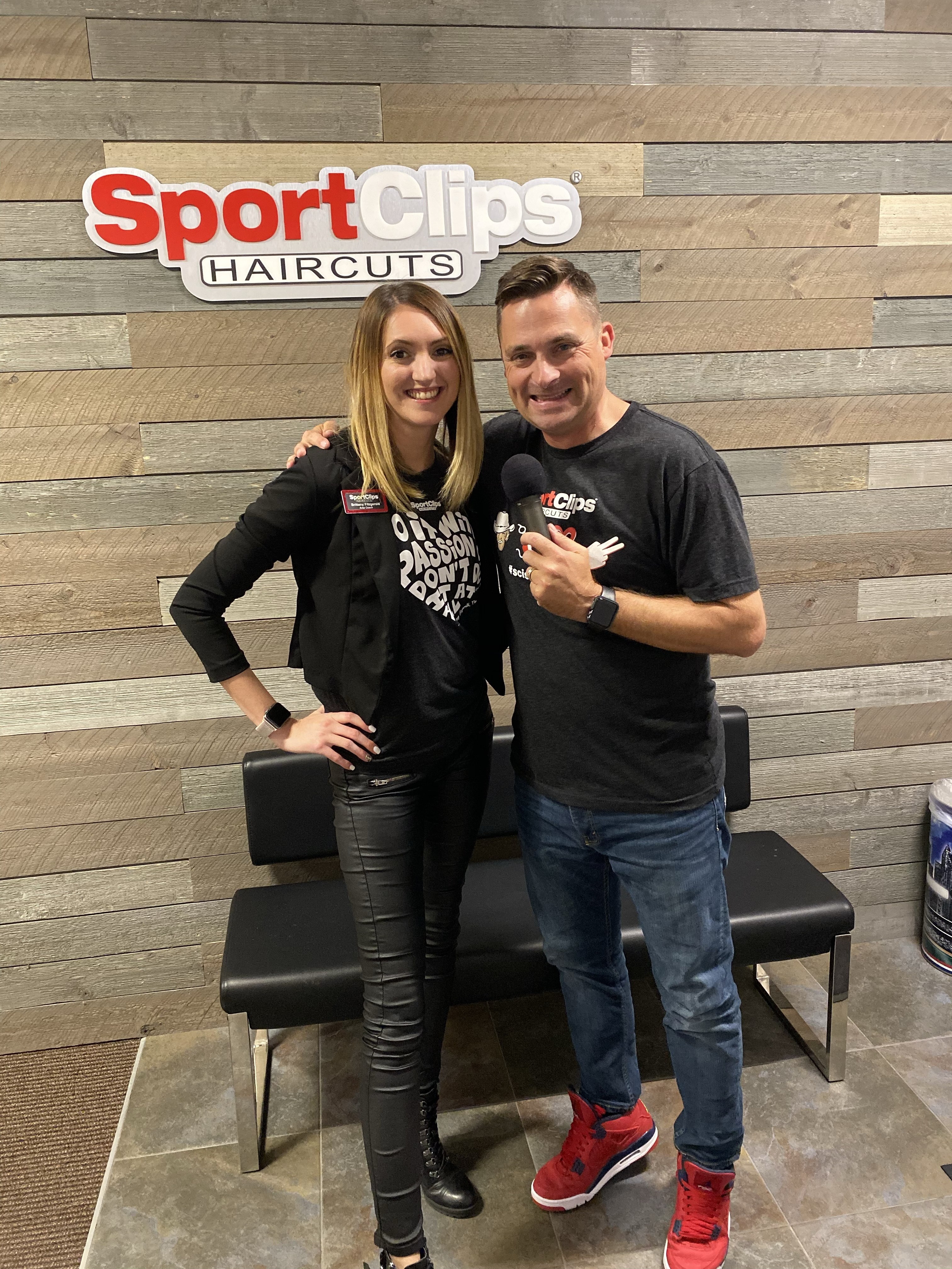 Man and woman standing in front of Sport Clips sign holding a microphone